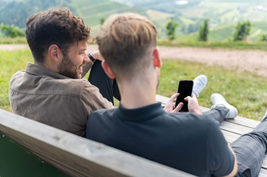 Zwei Männer sitzend auf einer Bank schauen gemeinsam auf ein Handy. Sitzen in einer grünen schönen Umgebung.