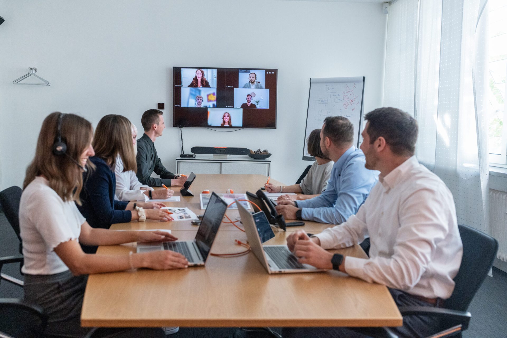 Meetingraum mit verschiedenen Personen die gleichzeitig in einem Onlinemeeting mit verschiedenen Personen sprechen. Es sind Laptops und Büromaterialien auf dem Tisch verteilt.
