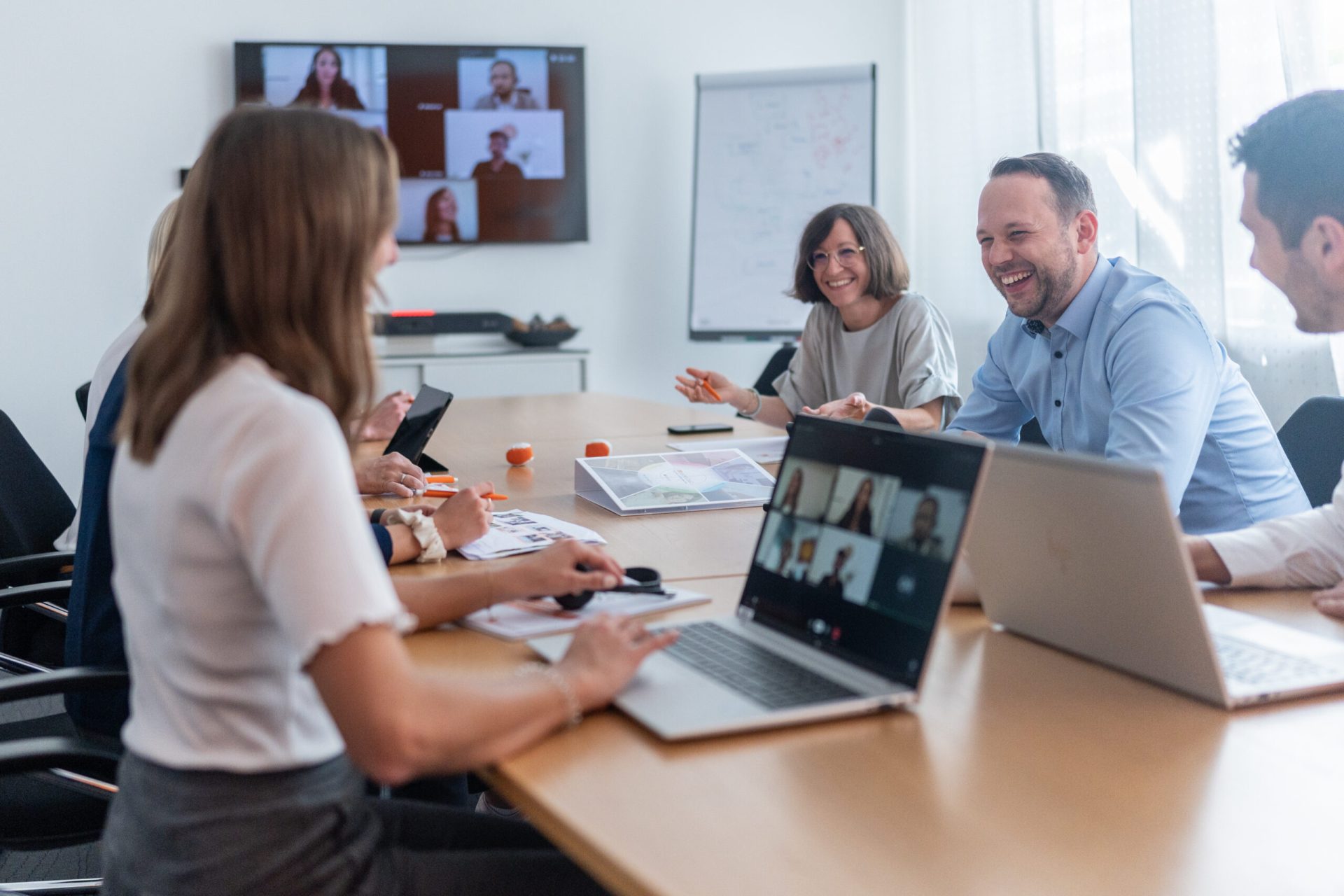 Mehrere Personen sind gemeinsam in einem Meeting in einem Meetingraum. Verschiedene Personen haben ihre Laptops dabei und es sind noch weitere Personen aus einem Onlinemeeting dazu geschalten. Sie lachen und diskutieren.