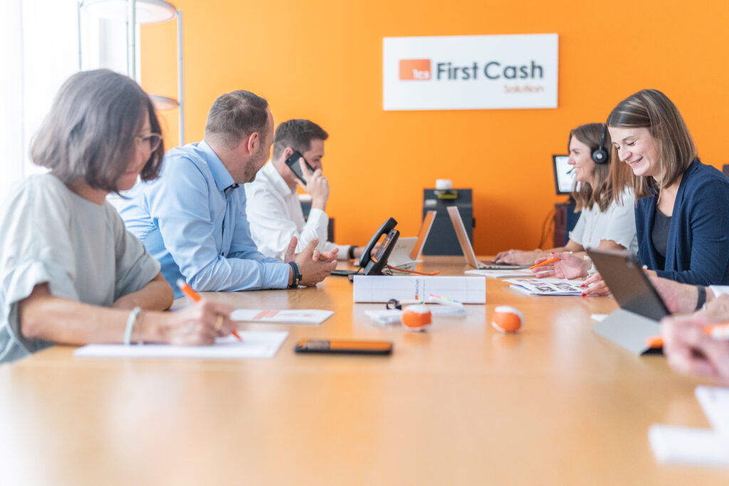 Mehrere Personen sitzen in einem Meetingraum mit einer orangenen Wand mit dem Logo der First Cash Solution GmbH. Eine Person trägt ein Headset und telefoniert, die anderen Personen erarbeiten verschiedene Themen.