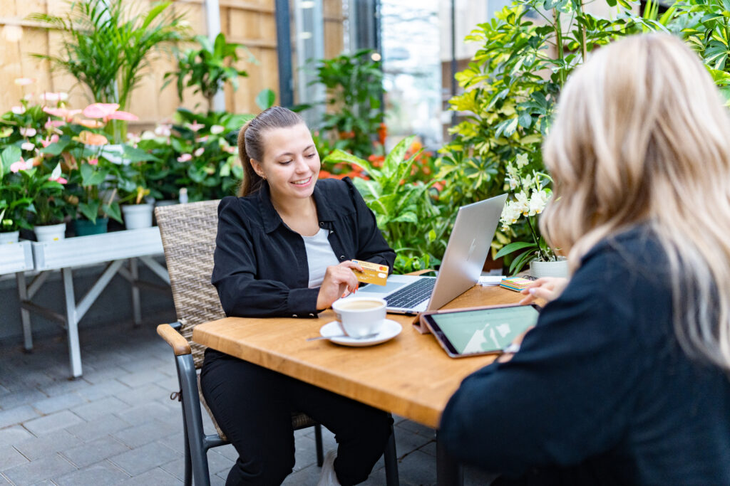 2 Frauen sitzen gemeinsam an einem Restaurant Tisch, umgeben von Pflanzen. Vor einer lächelnden Frau steht ein Laptop, daneben eine Tasse Tee und sie hält eine Kreditkarte in der Hand. Die Frau ihr gegenüber zeigt ihr etwas am Ipad.